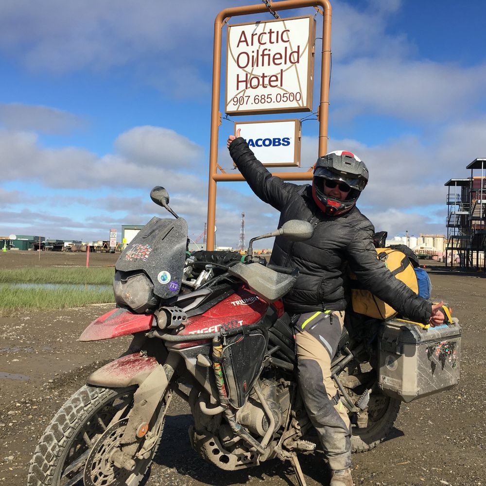 Caroline in helmet and riding jacket, thumbs up, sitting on Boo, a mud-covered red Triumph Tiger 800, under the Arctic Oilfield Hotel sign in Deadhorse, Alaska