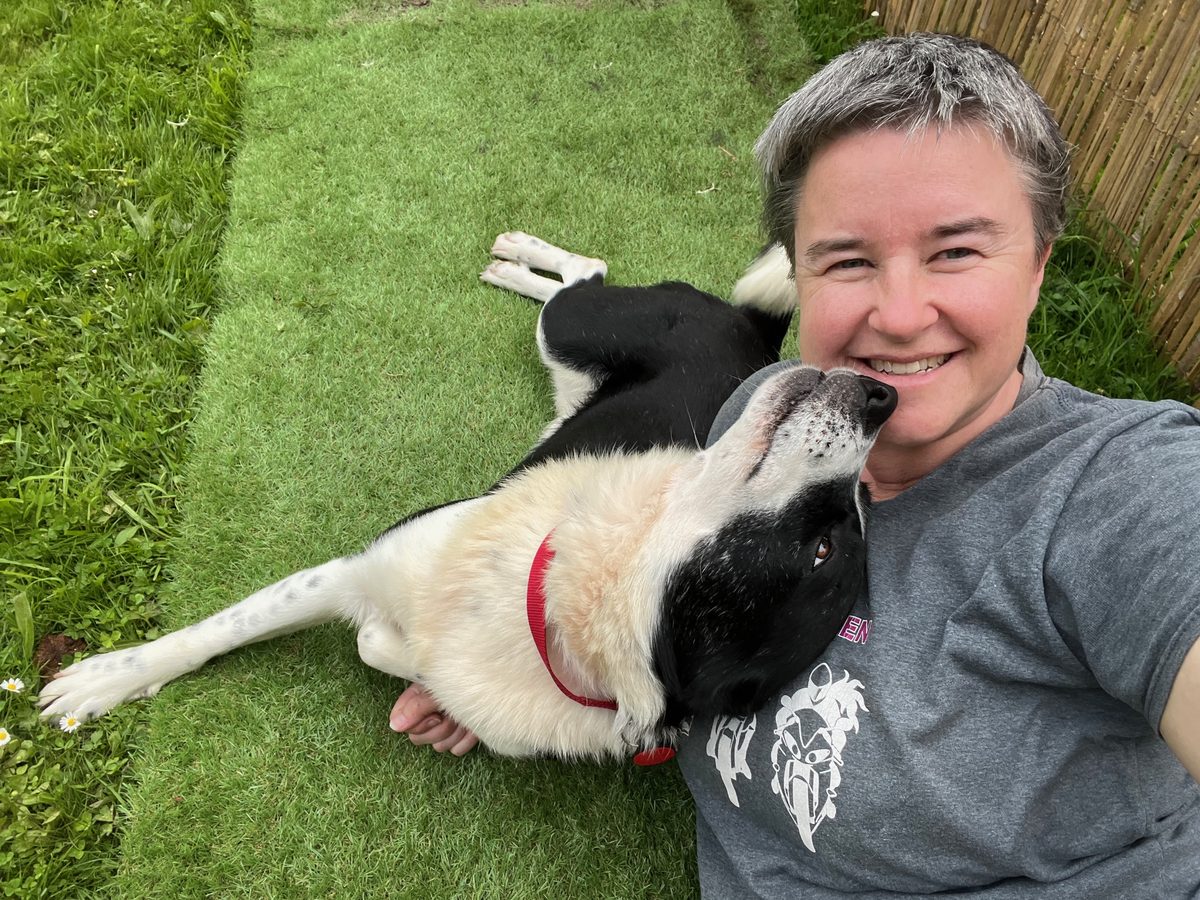 Caroline smiling in a selfie on the grass, with Tex, a black-and-white foster dog, lying relaxed beside her